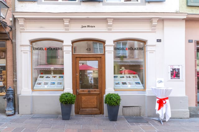 Bureau immobilier Engel & Völkers avec une porte en bois, de grandes fenêtres et des plantes en pot sur un trottoir en briques.