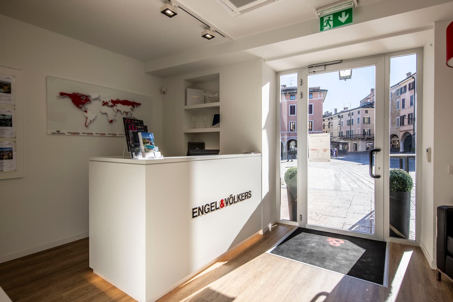 Engel & Völkers office interior with a white reception desk, world map, and open door to a European town square.