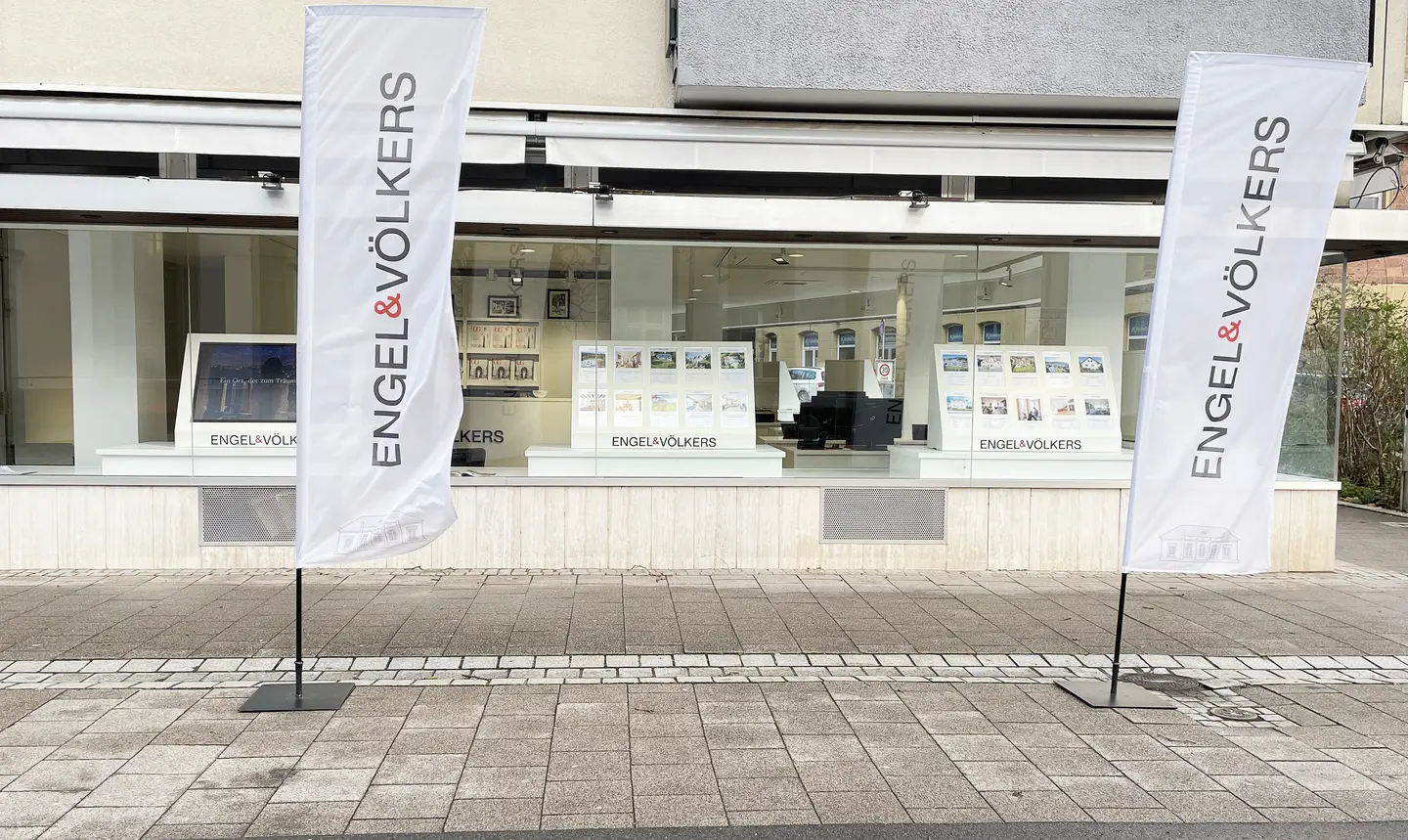Engel & Völkers real estate office front with white flags on a paved sidewalk. The flags display the company name in gray and red.