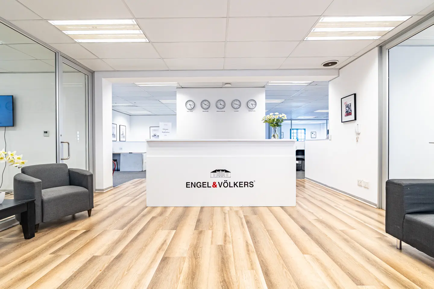 Engel & Völkers office interior with a white reception desk, world clocks, gray armchairs, and light wood floors.