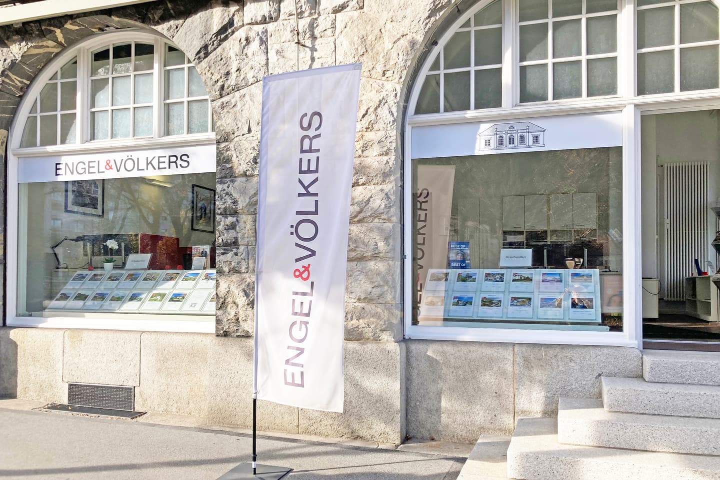 Engel & Völkers real estate office exterior with stone facade, arched windows, and a white flag with the company logo.