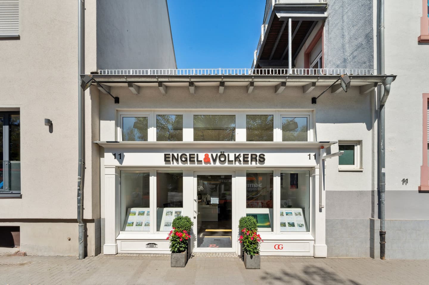 Exterior of Engel & Völkers real estate office with white trim, large windows, and potted red flowers.