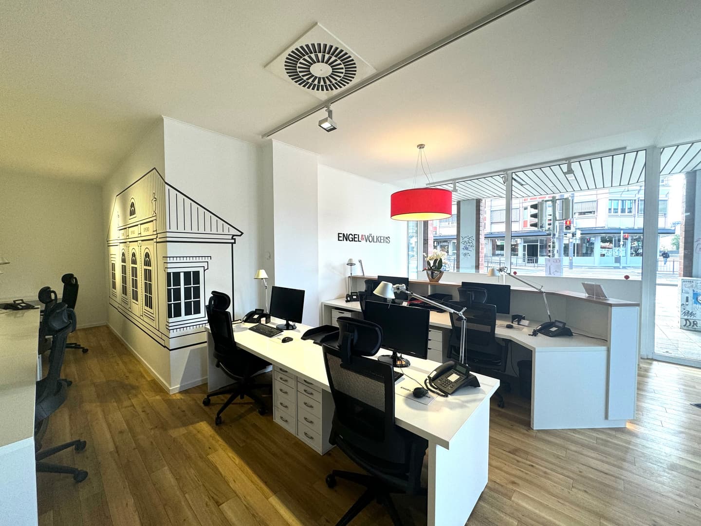 Engel & Völkers Büro-Interieur mit weißen Schreibtischen, schwarzen Stühlen, Computern und einem Wandgemälde eines Gebäudes an der Wand. Eine rote Lampe hängt von der Decke.