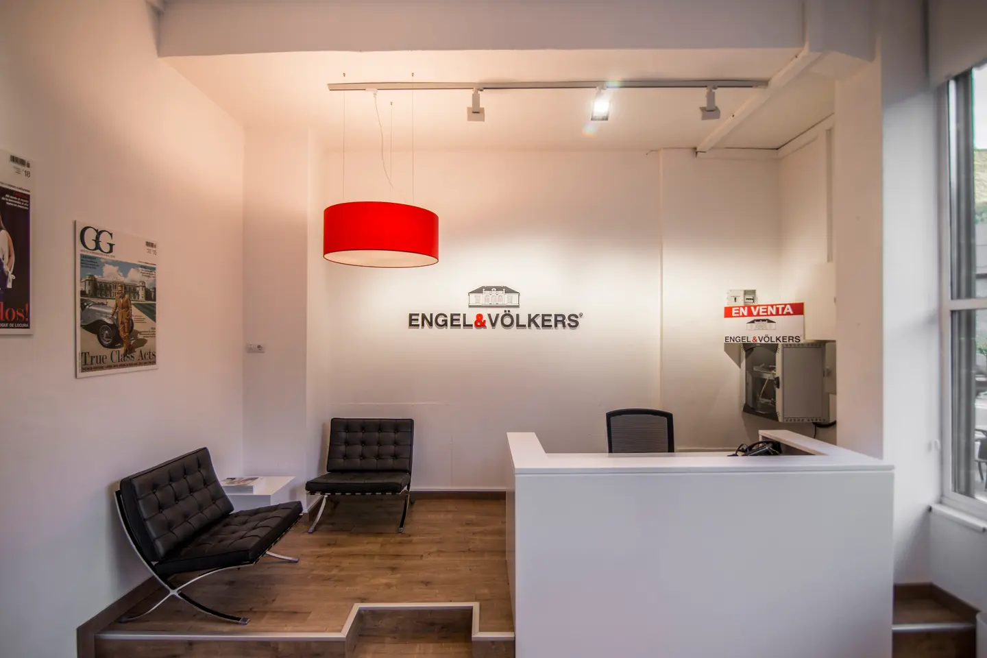 Oficina Engel & Völkers amb un taulell de recepció blanc, cadires negres i una làmpada penjant vermella. Es veu el cartell "En Venta".