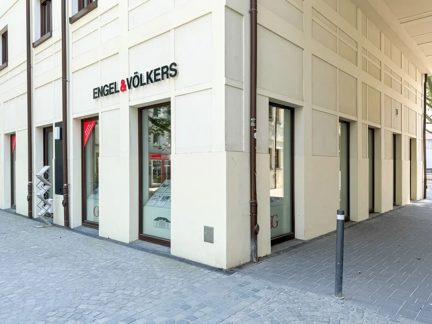Engel & Völkers real estate office on a corner. The building is white with dark-framed windows. A gray cobblestone street is in front.