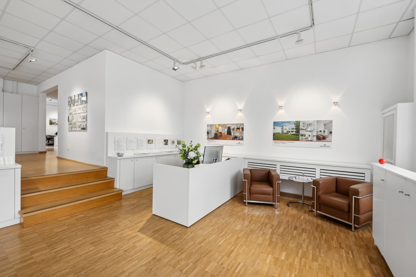 Engel & Völkers Immobilienbüro mit weißer Fassade, Glasfenstern mit Immobilieneinträgen und schwarzen Pflanzgefäßen mit grünen Büschen.