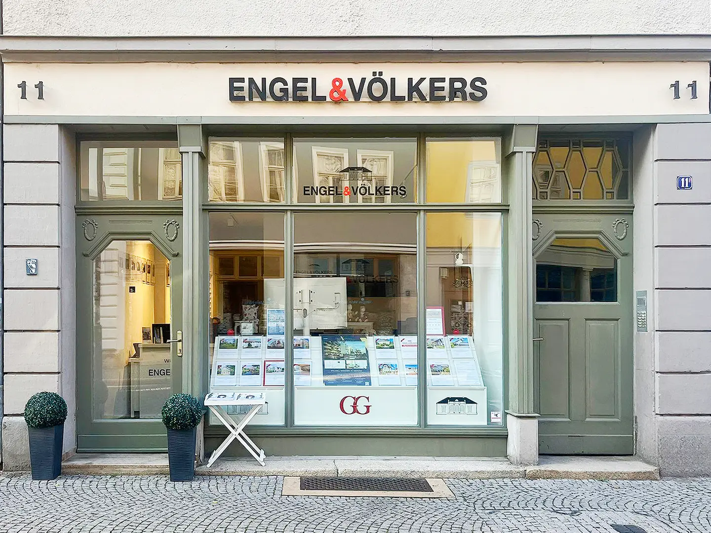 Engel & Völkers Immobilienbüro mit einer großen Fensterfront mit Immobiliendarstellungen und einer grünen Tür.