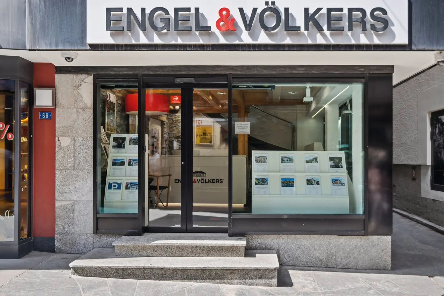 Vista esterna dell'ufficio immobiliare Engel & Völkers con porte in vetro, annunci immobiliari e gradini in pietra grigia.