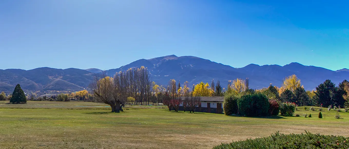 Vista panoràmica d'un camp verd amb arbres, un edifici i muntanyes sota un cel blau clar.