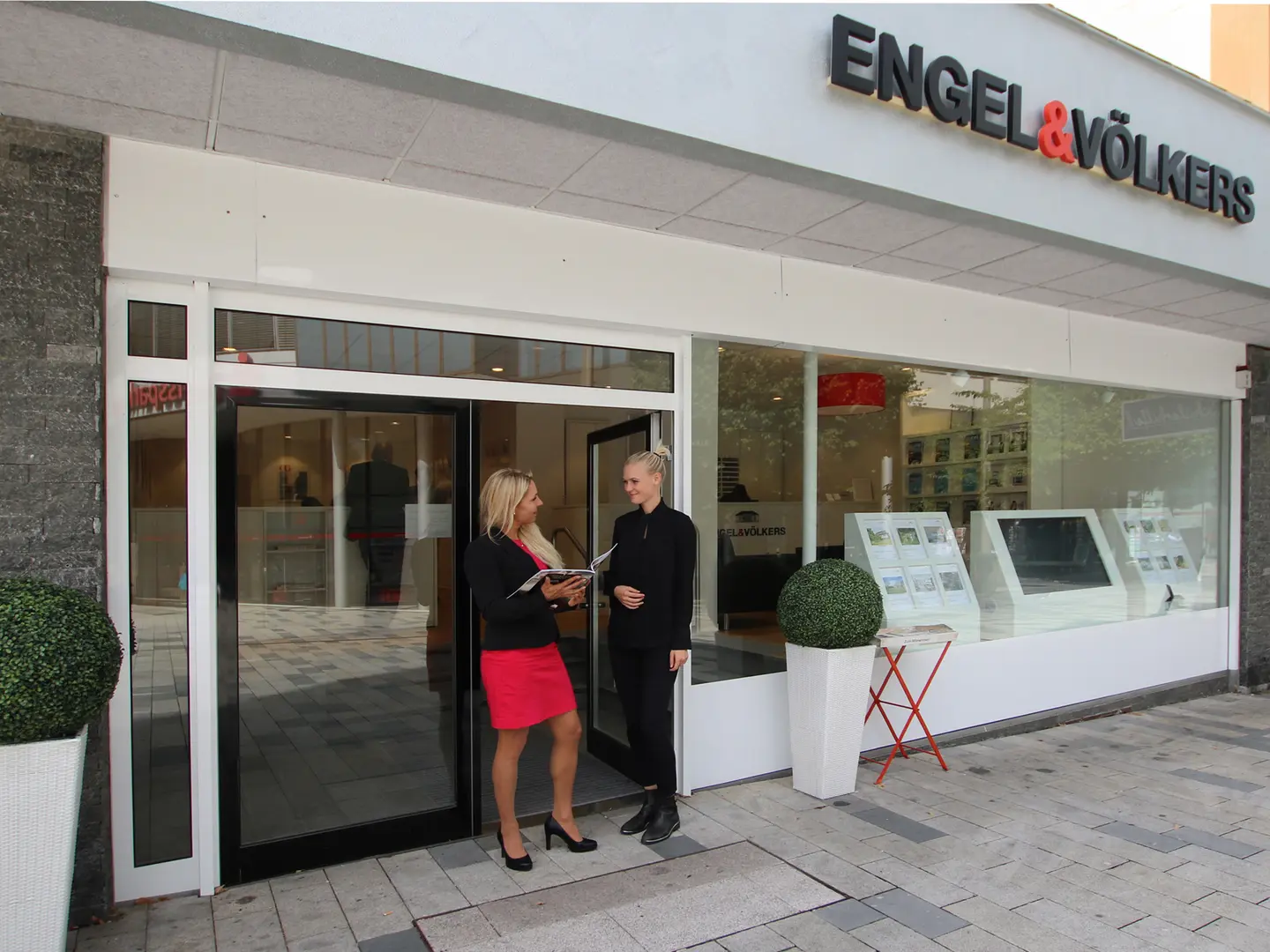 Zwei Frauen stehen vor einem Engel & Völkers Immobilienbüro. Eine im roten Kleid hält eine Broschüre, die andere ist schwarz gekleidet.