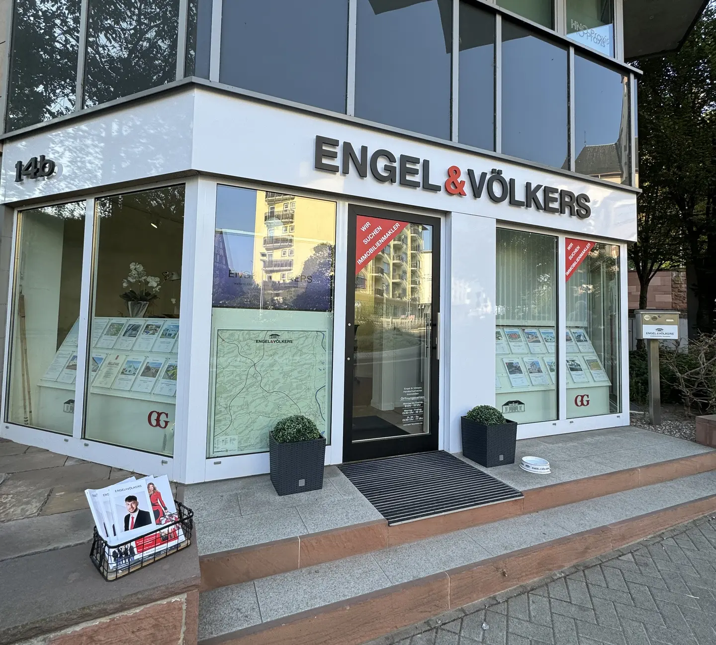 Engel & Völkers real estate office with glass windows and door. A black basket with brochures sits on the steps. Address number 14b is visible.