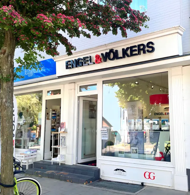 Engel & Völkers Immobilienbüro mit weißer Verkleidung und schwarzer Beschriftung. Links steht ein Baum mit rosa Blüten.