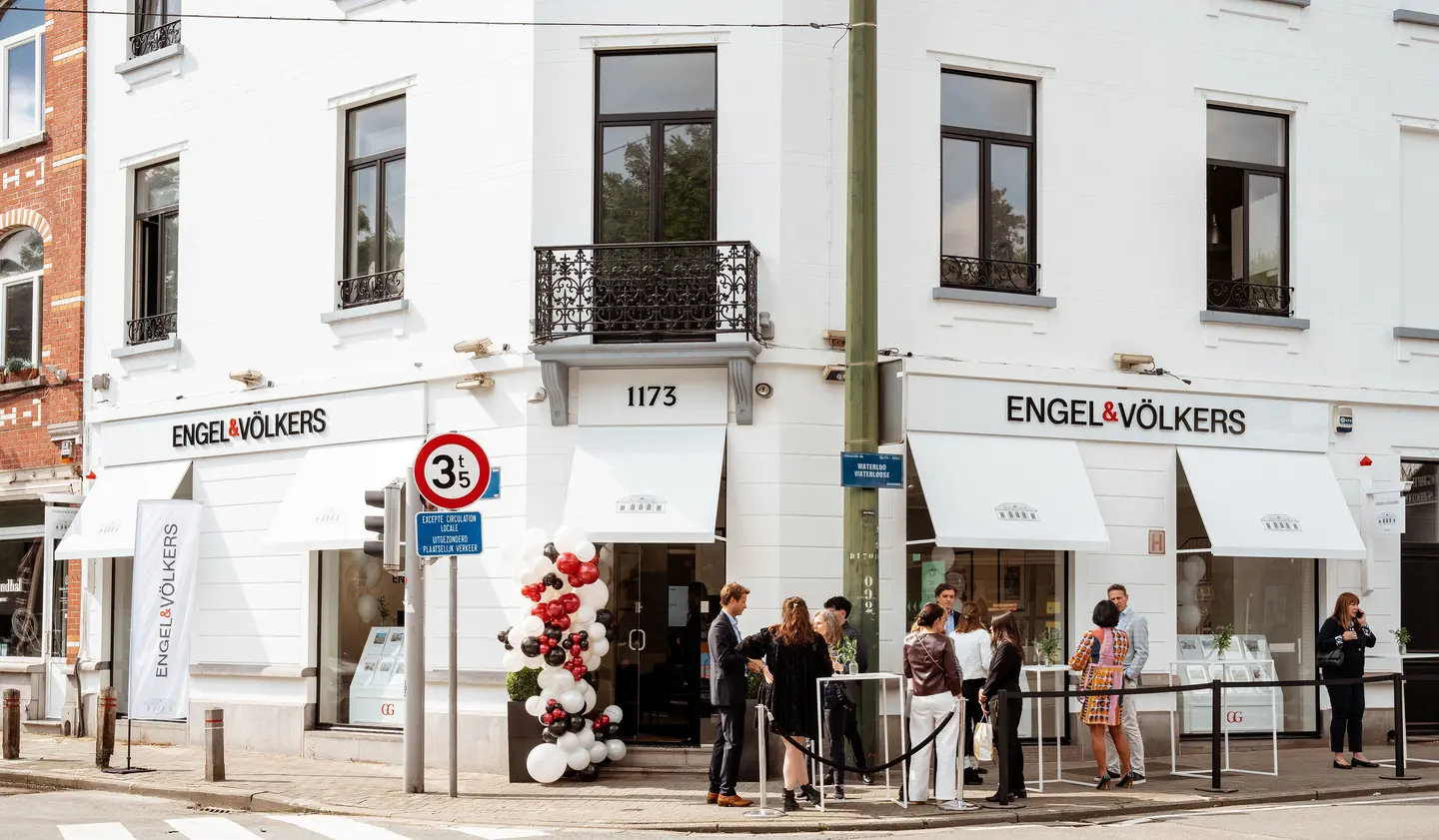 Engel & Völkers office exterior with people gathered outside. White building with black window frames and awnings. Balloons decorate the entrance.