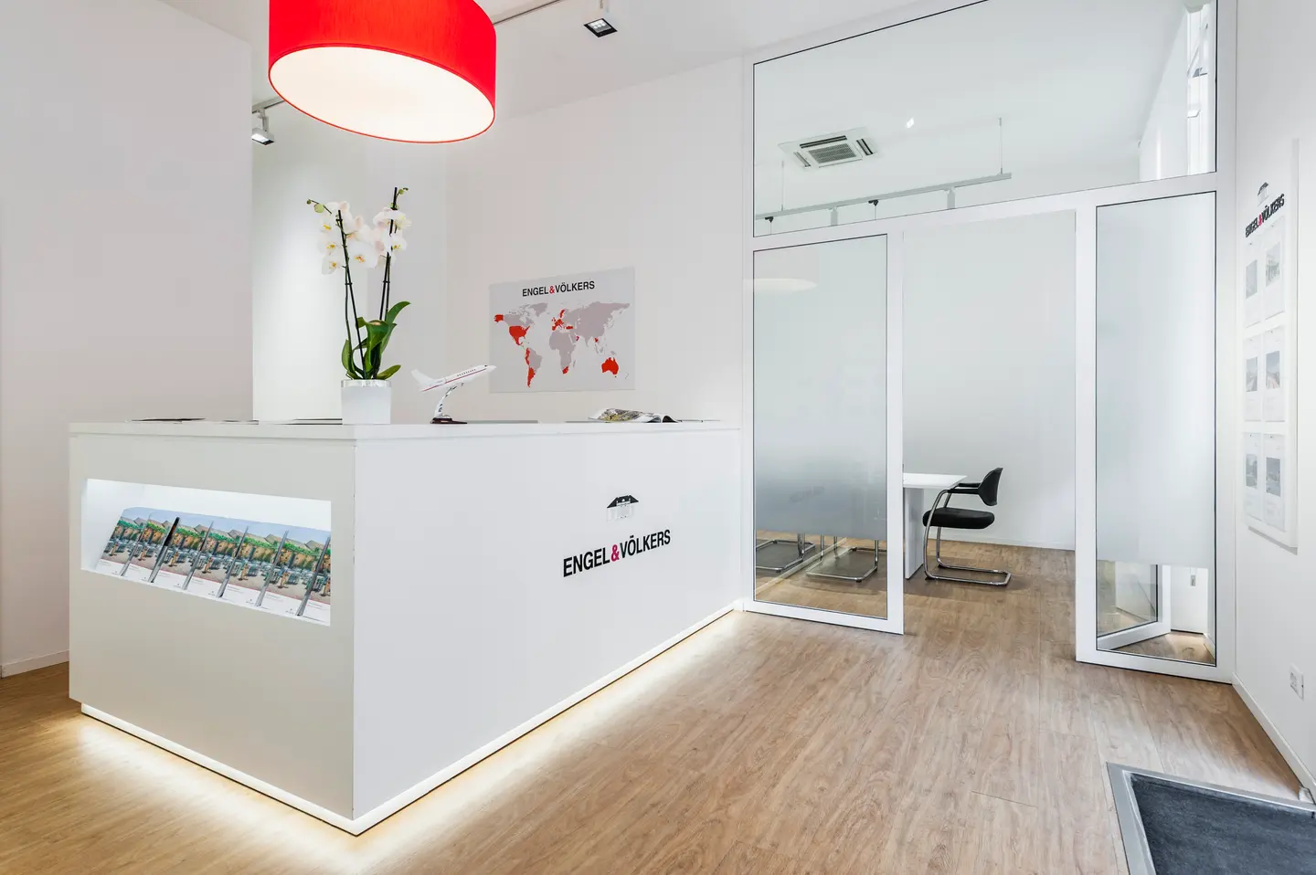 Engel & Völkers Empfangsbereich mit weißem Schreibtisch, roter Lampe, Weltkarte und Büro mit Glaswänden.