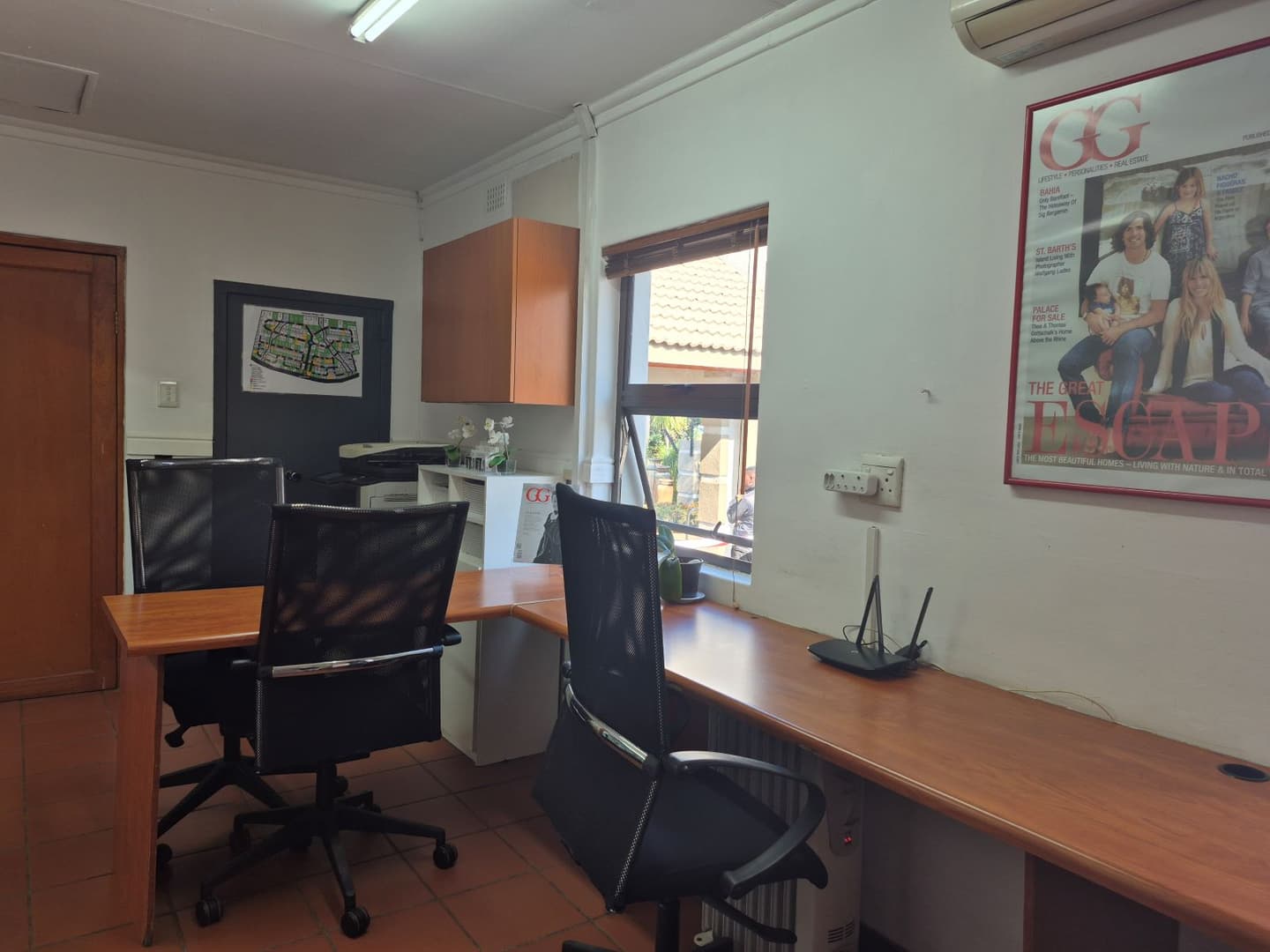 Office interior with wooden desks, black mesh chairs, and terracotta tile floor. A window overlooks a building. A real estate magazine hangs on the wall.