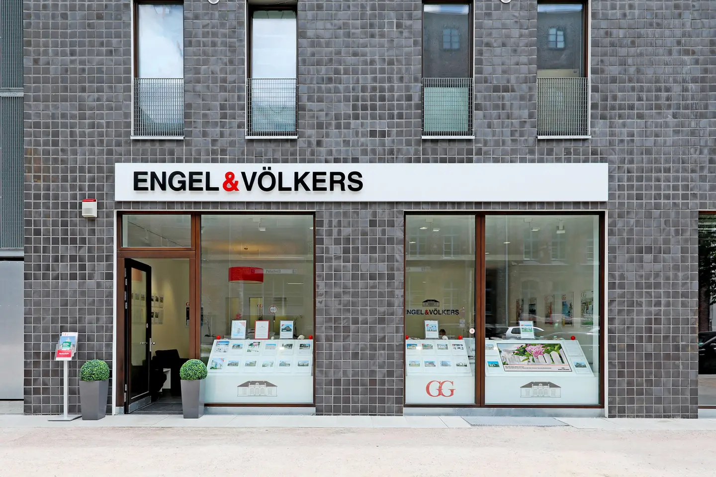 Engel & Völkers Immobilienbüro mit einer grauen Fliesen Fassade, großen Fenstern, die Immobilienangebote zeigen, und einer offenen Tür.