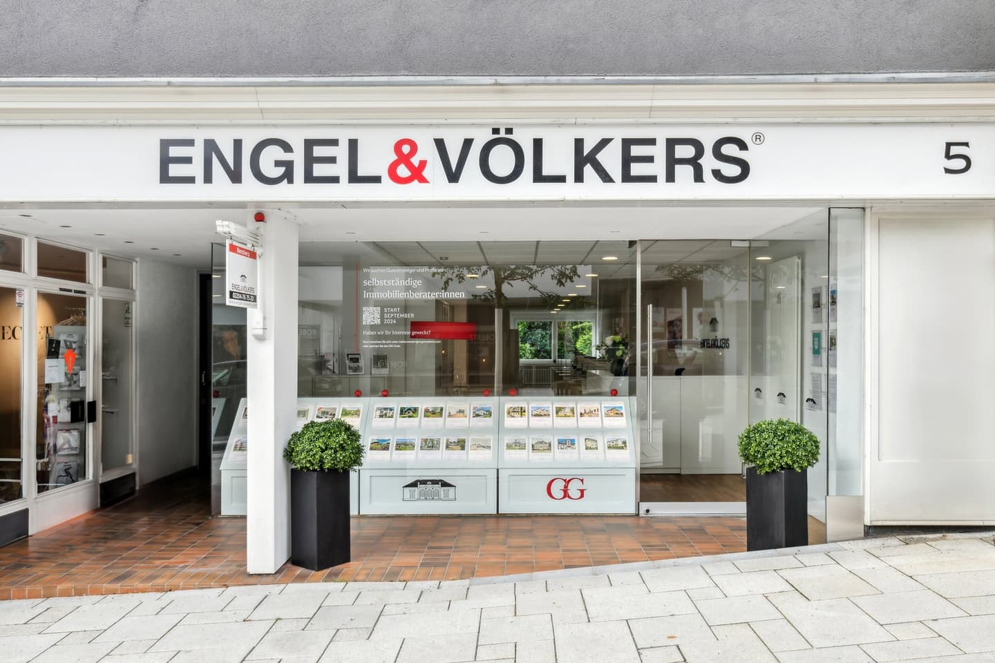 Engel & Völkers real estate office storefront with glass windows, property listings, and potted plants on a brick walkway.