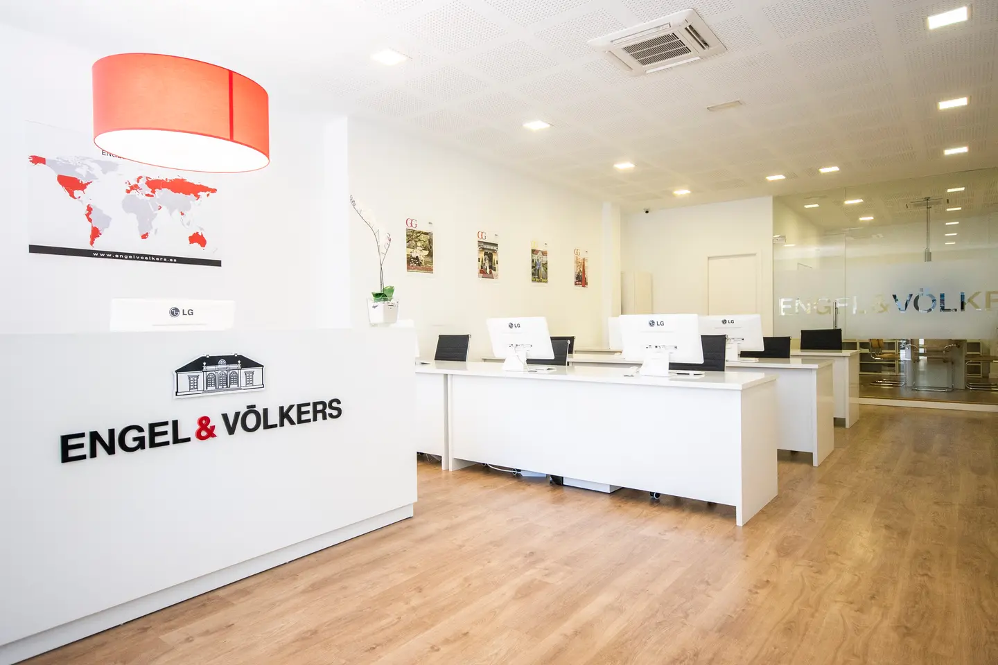 Engel & Völkers Bürointerieur mit weißen Schreibtischen, LG-Monitoren und einer roten Lampe. Der Boden ist aus hellem Holz.