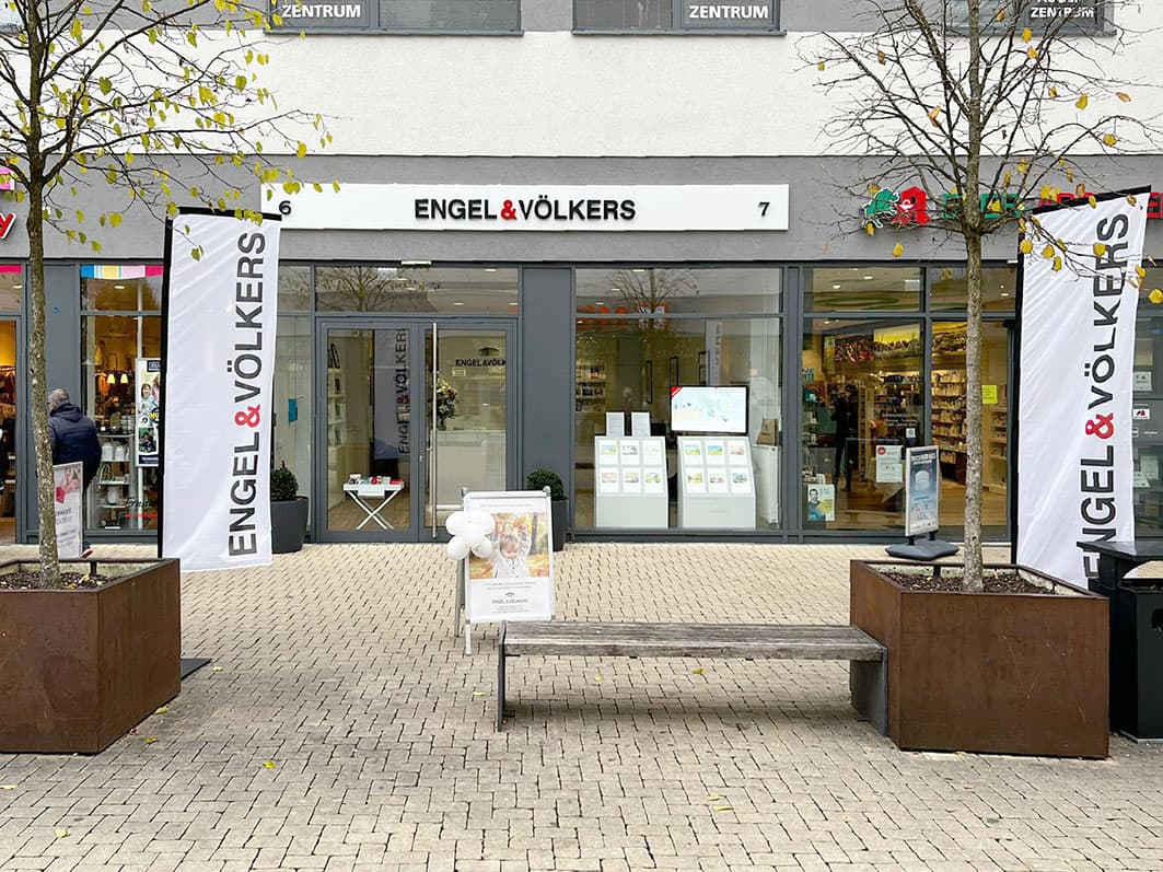 Engel & Völkers real estate office with glass storefront and flags on a brick walkway with a bench and trees in planters.