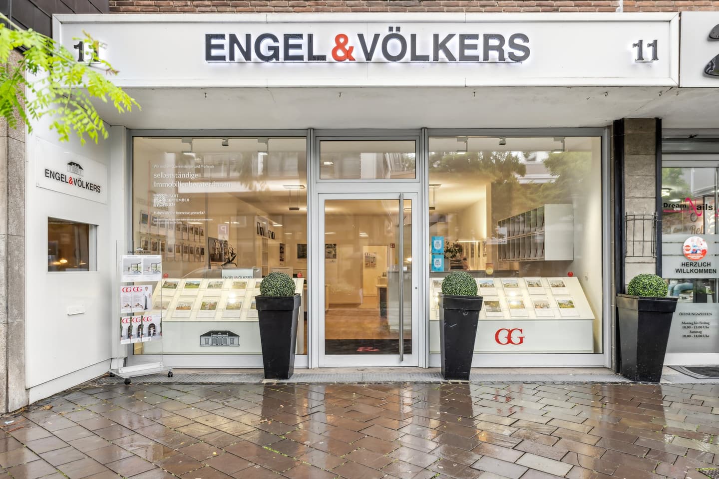 Engel & Völkers real estate office storefront with glass windows and door. Black planters flank the entrance. Magazines are displayed outside.