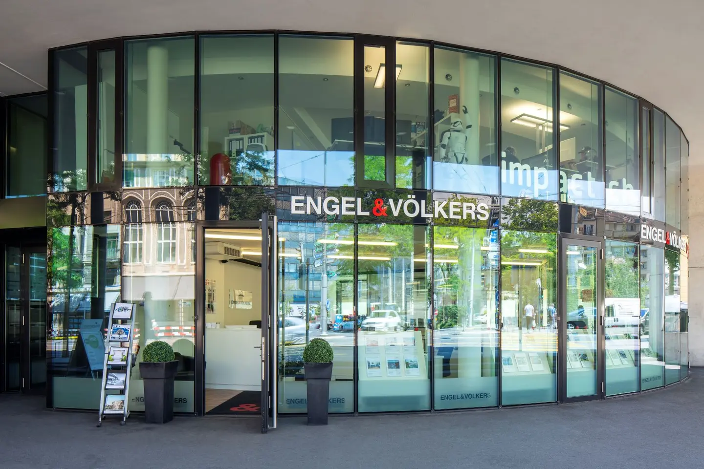 Engel & Völkers Immobilienbüro mit einer gebogenen Glasfassade. Das Logo ist über dem Eingang angebracht. Innenansicht des Empfangsbereichs.
