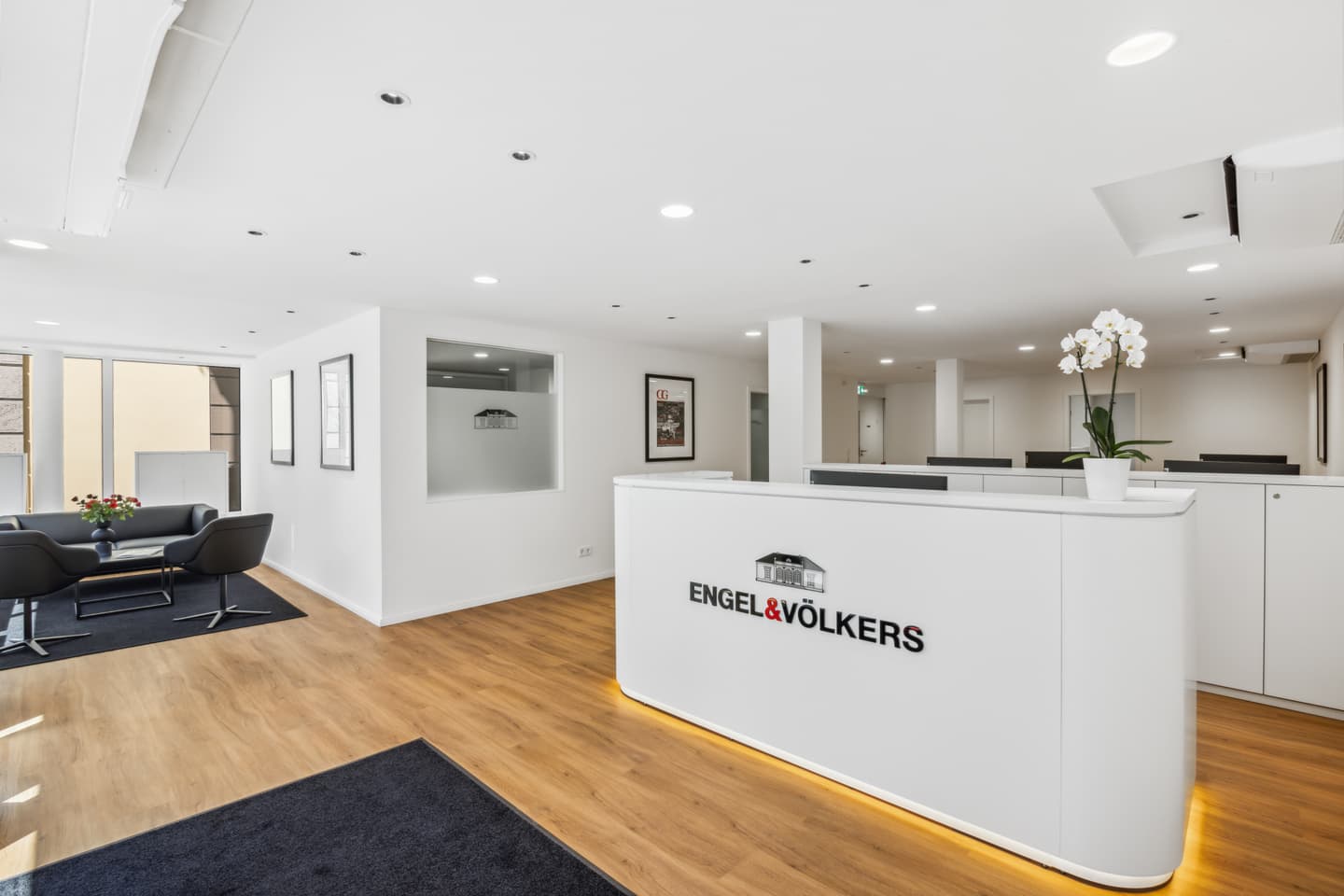 Helles, modernes Immobilienbüro mit weißen Wänden, Holzböden und einem Empfangstresen mit dem Engel & Völkers Logo. Ein Sitzbereich ist sichtbar.