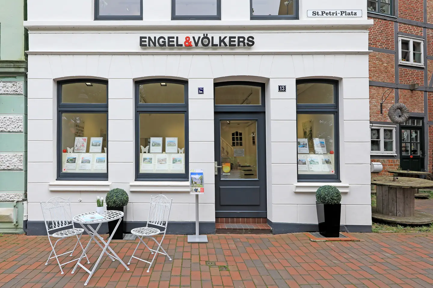 Exterior of Engel & Völkers real estate office with white facade, dark gray door and windows. A table and chairs sit on a brick walkway.