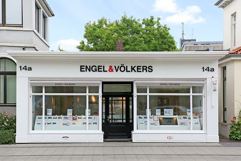 Außenansicht eines Engel & Völkers Immobilienbüros mit weißen Zierleisten, schwarzer Tür und Schaufensterauslagen.