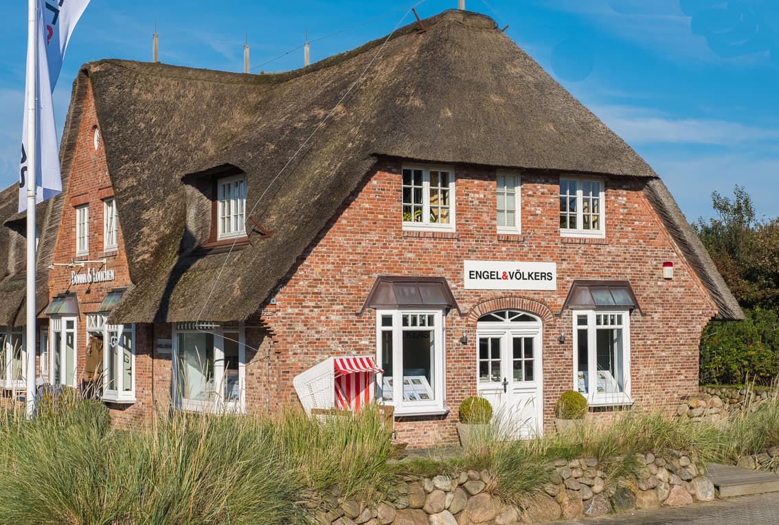 Engel & Völkers real estate office in a brick building with a thatched roof. Tall grass and a beach chair are in front.
