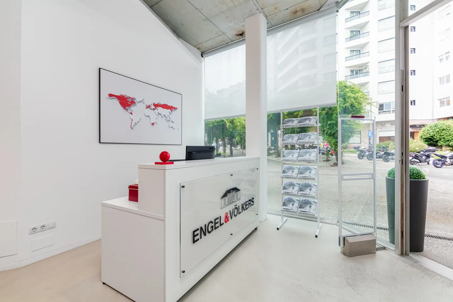 Bright, modern real estate office with white reception desk, Engel & Völkers logo, world map art, and brochure rack by a large window.