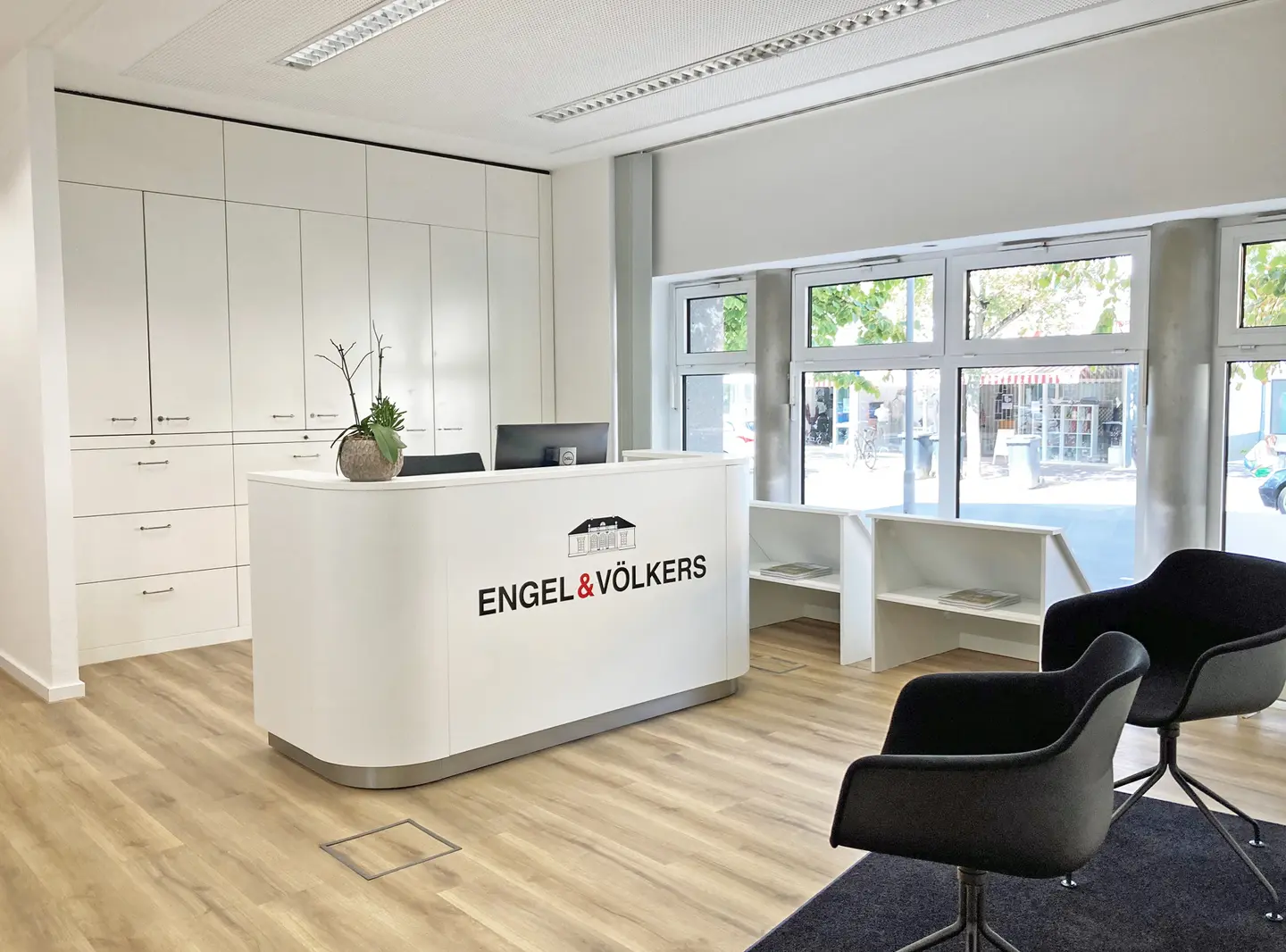 Helles Immobilienbüro mit weißem Empfangstresen, Engel & Völkers Logo, schwarzen Stühlen und hellen Holzböden. Große Fenster bieten Straßenblick.