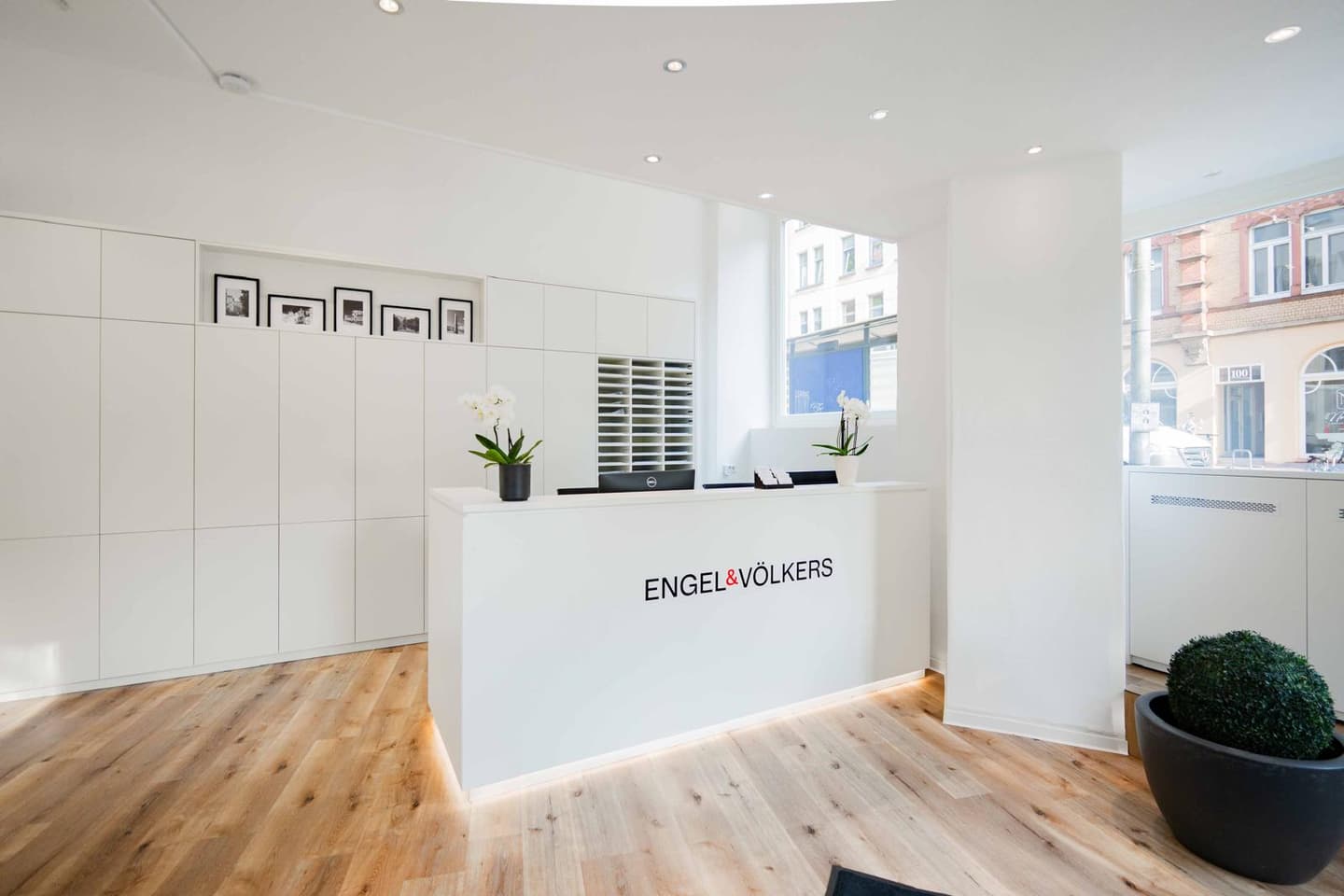 Engel & Völkers Bürointerieur mit einem weißen Empfangstresen, Holzböden und weißen Wänden. Das Firmenlogo befindet sich auf dem Tresen.