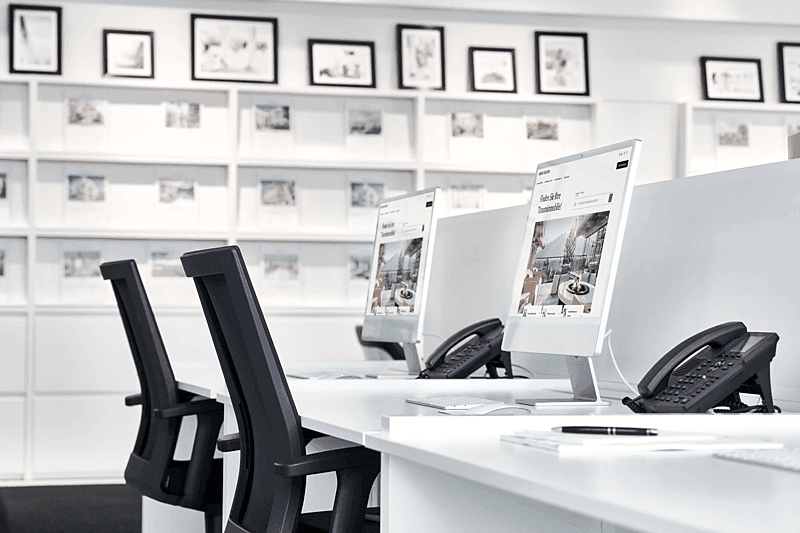 A bright real estate office with white desks, black chairs, computers, and phones. A wall of framed property photos is in the background.