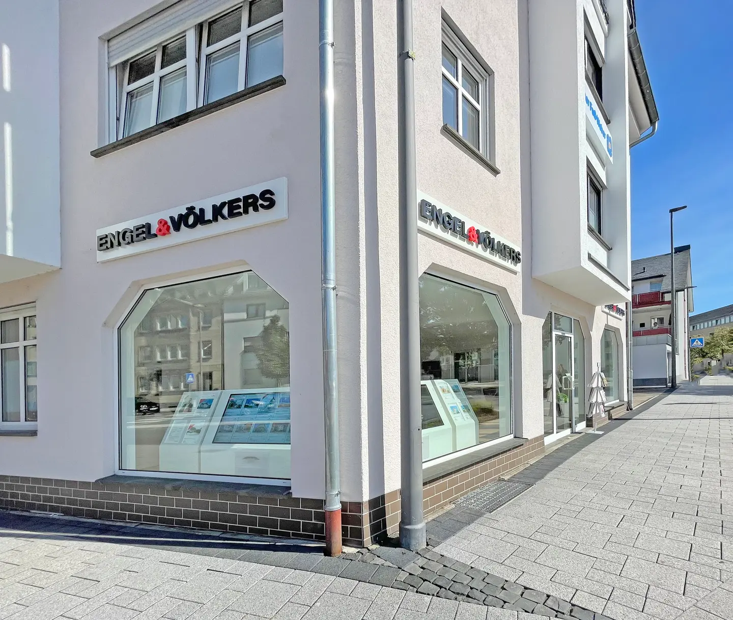 Engel & Völkers real estate office exterior. White building with large windows displaying property listings. Paved sidewalk in front.