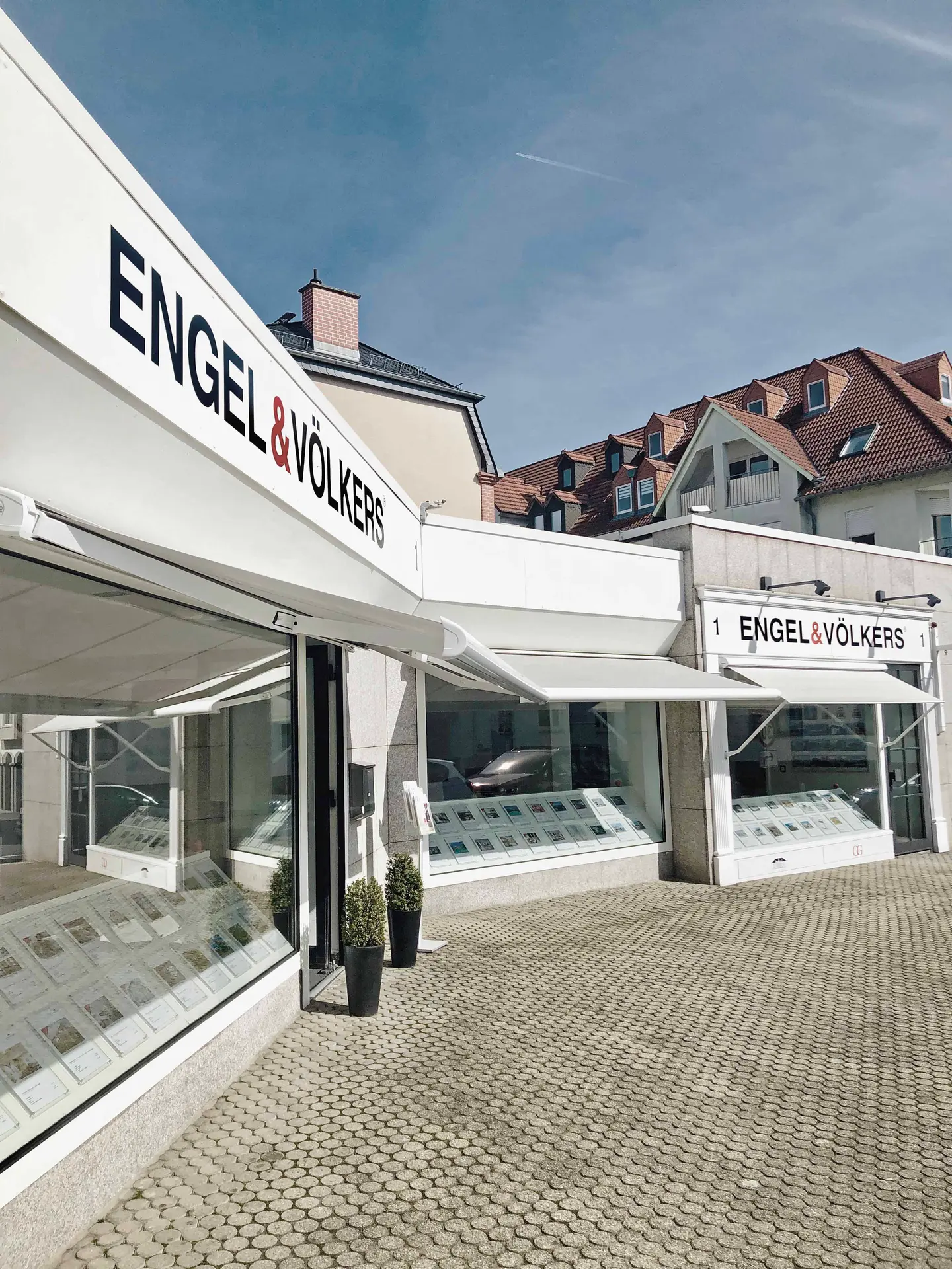 Engel & Völkers real estate office exterior with white awnings, display windows, and gray cobblestone walkway.