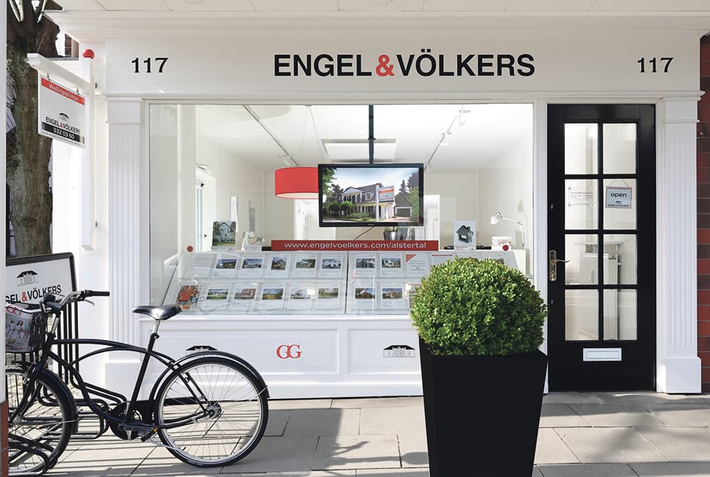 Engel & Völkers Immobilien-Schaufenster mit einem schwarzen Fahrrad, das draußen geparkt ist. Ein Fernseher zeigt ein Haus zum Verkauf im Fenster.