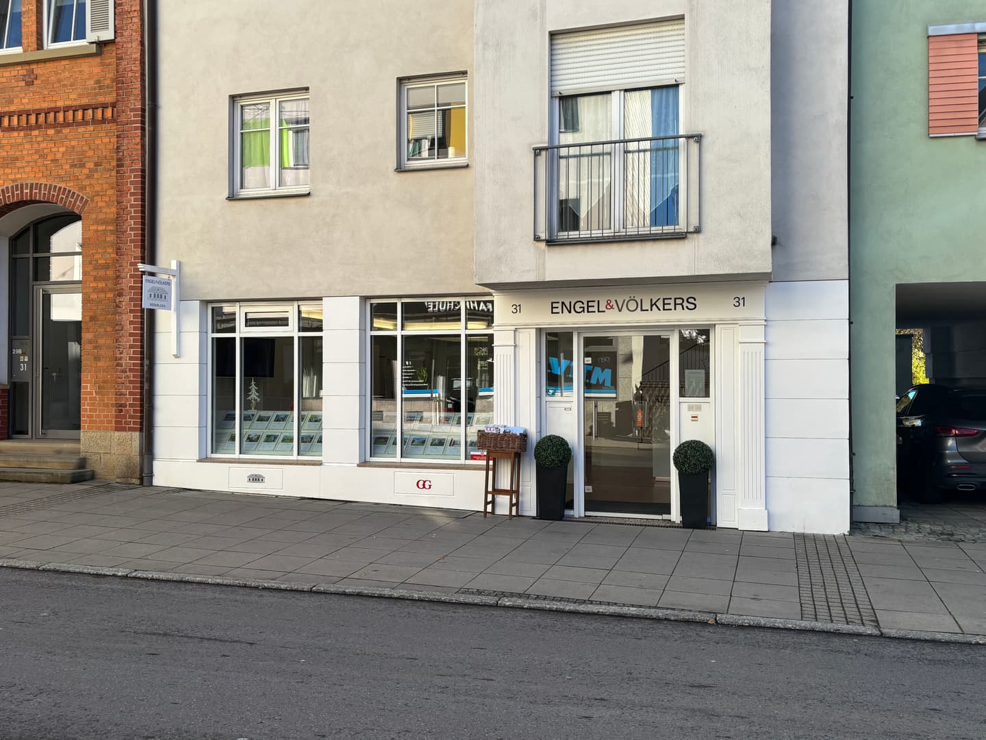 Engel & Völkers real estate office with a white facade, large windows displaying property listings, and potted plants flanking the entrance.