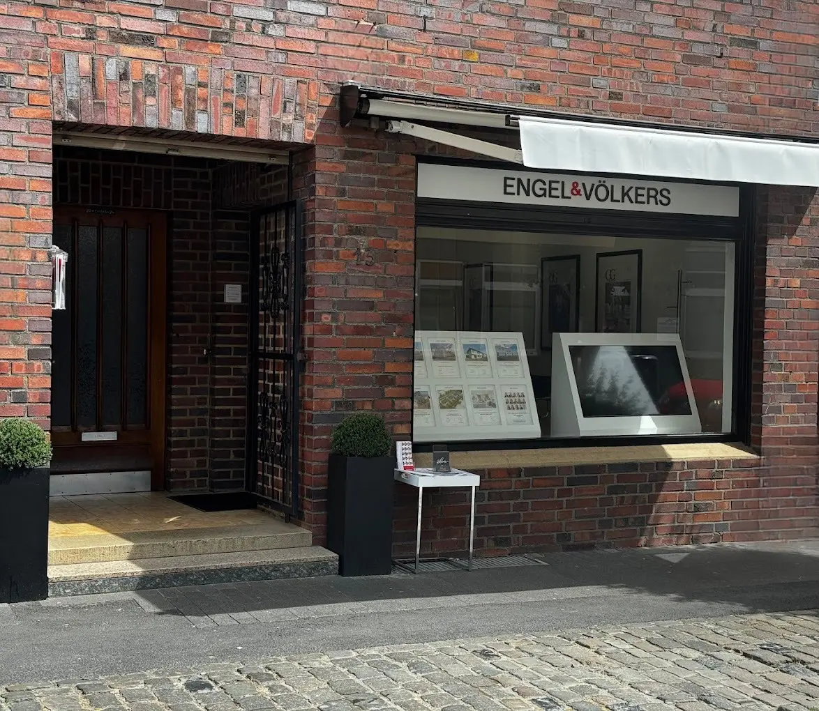 Exterior of an Engel & Völkers real estate office in a red brick building with a white awning and window display.