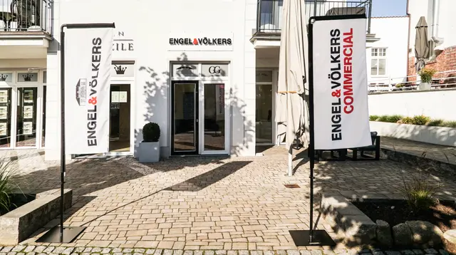Engel & Völkers real estate office with white facade, brick walkway, and branded flags on a sunny day.