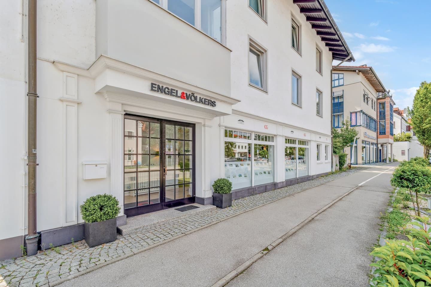 Engel & Völkers Immobilienbüro mit weißer Fassade, schwarz gerahmten Fenstern und Immobiliendisplays. Zwei Topfpflanzen flankieren den Eingang.