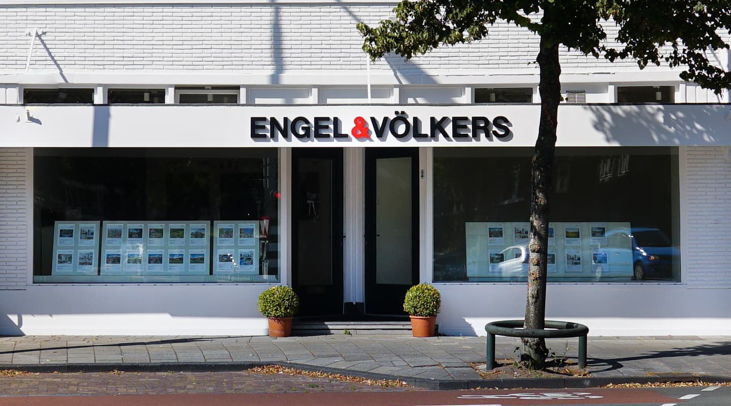 Engel & Völkers real estate office with a white brick facade and large windows displaying property listings. A tree stands in front.