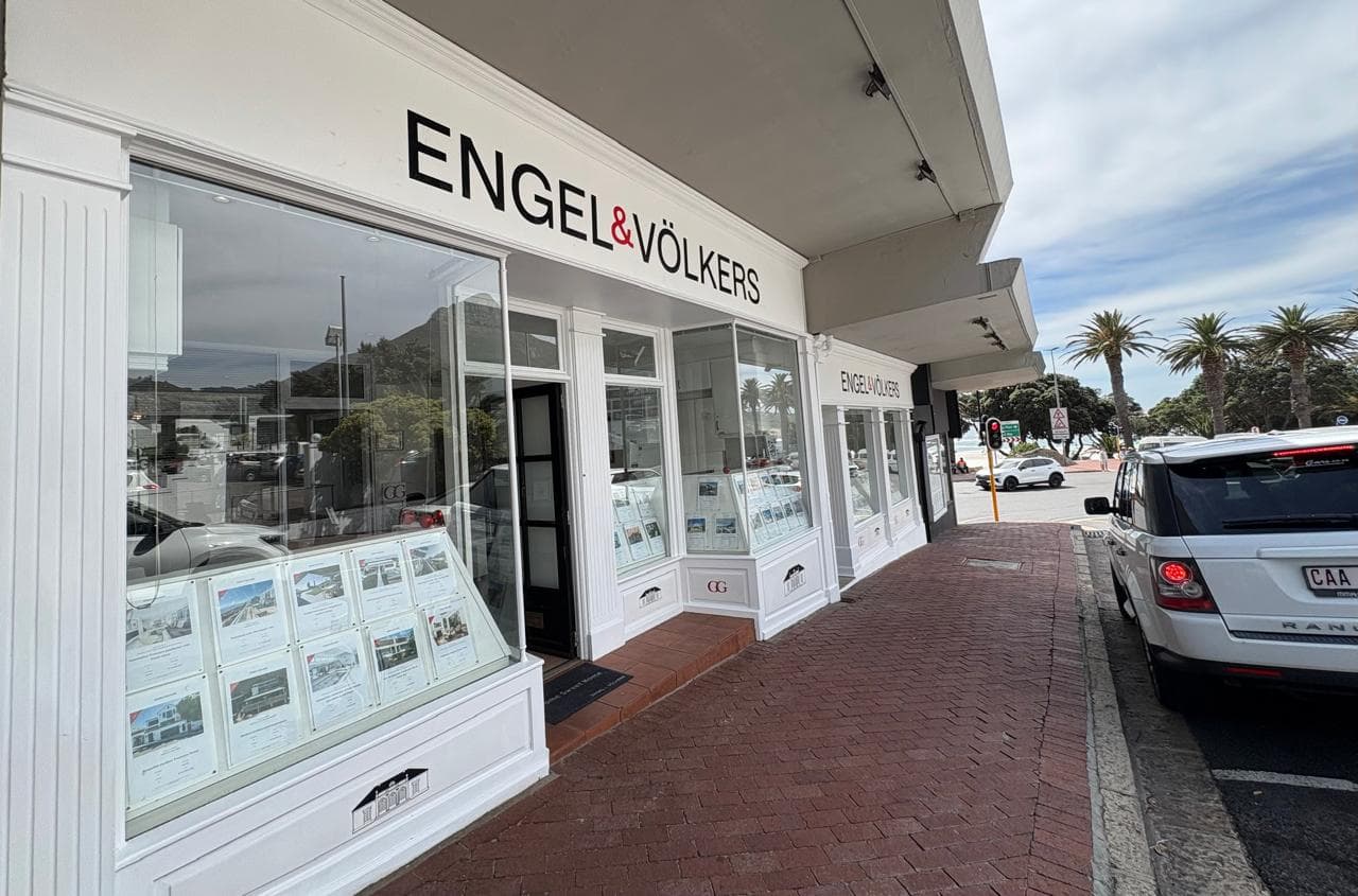 Engel & Völkers real estate office with white trim and brick sidewalk. Palm trees and a white SUV are visible in the background.