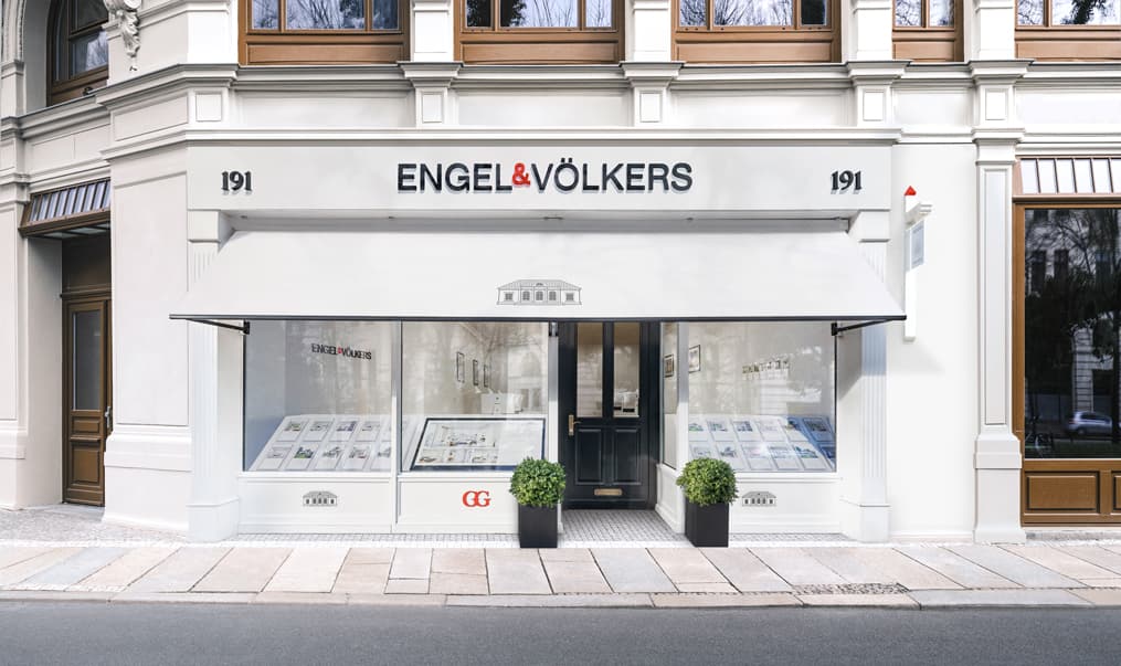 Engel & Völkers real estate office exterior. White building with a black door, awning, and window displays. Two potted plants flank the entrance.