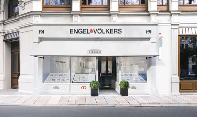 Ufficio immobiliare Engel & Völkers, esterno. Edificio bianco con porta, tenda e vetrine nere. Due piante in vaso fiancheggiano l'ingresso.