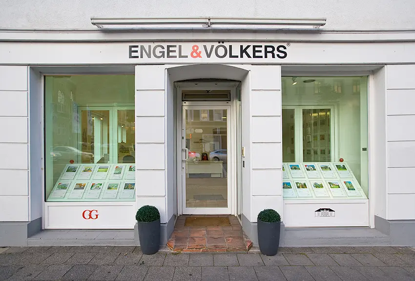 Engel & Völkers real estate office storefront with property listings in the windows and potted plants flanking the entrance.