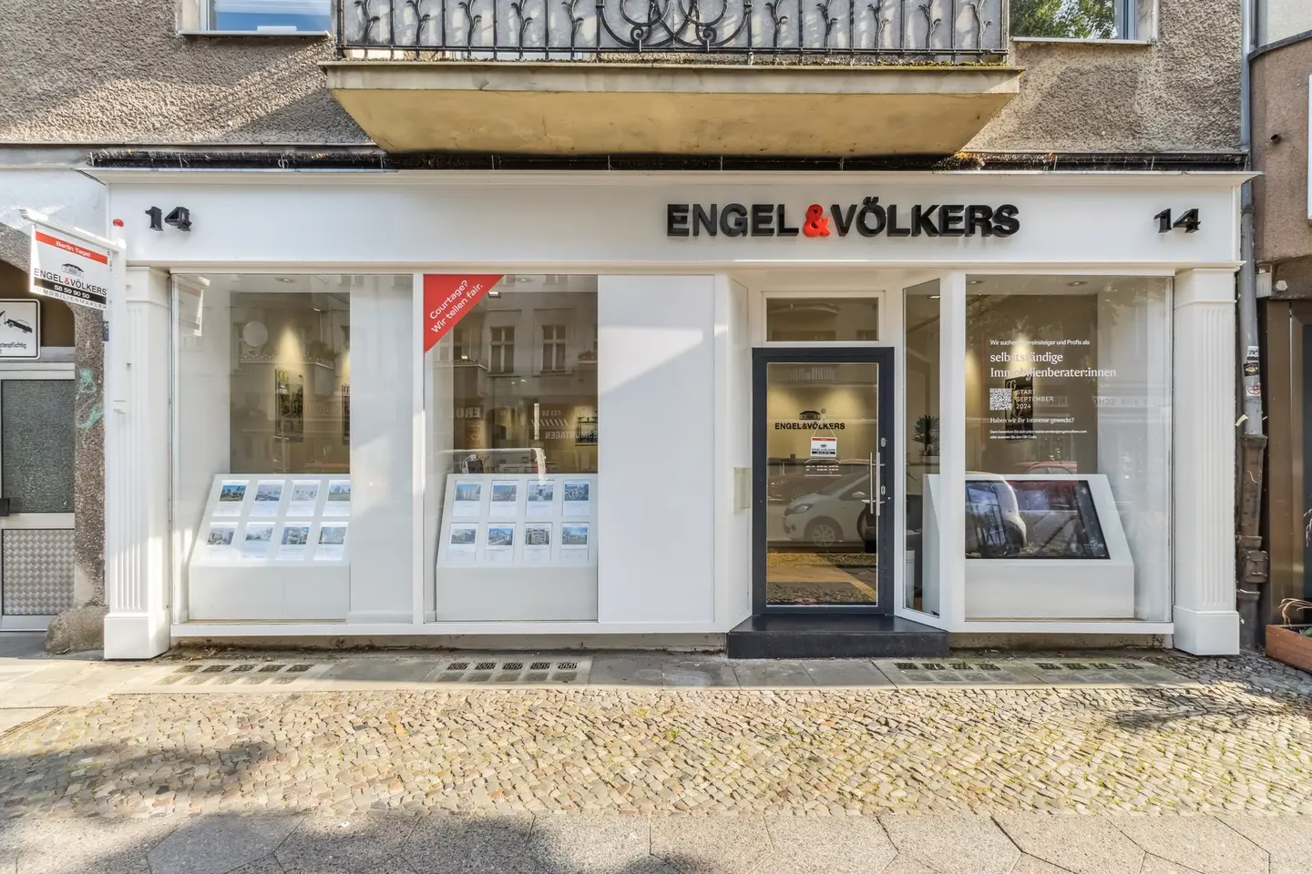 Engel & Völkers real estate office storefront with white trim and large windows displaying property listings. Cobblestone sidewalk in front.