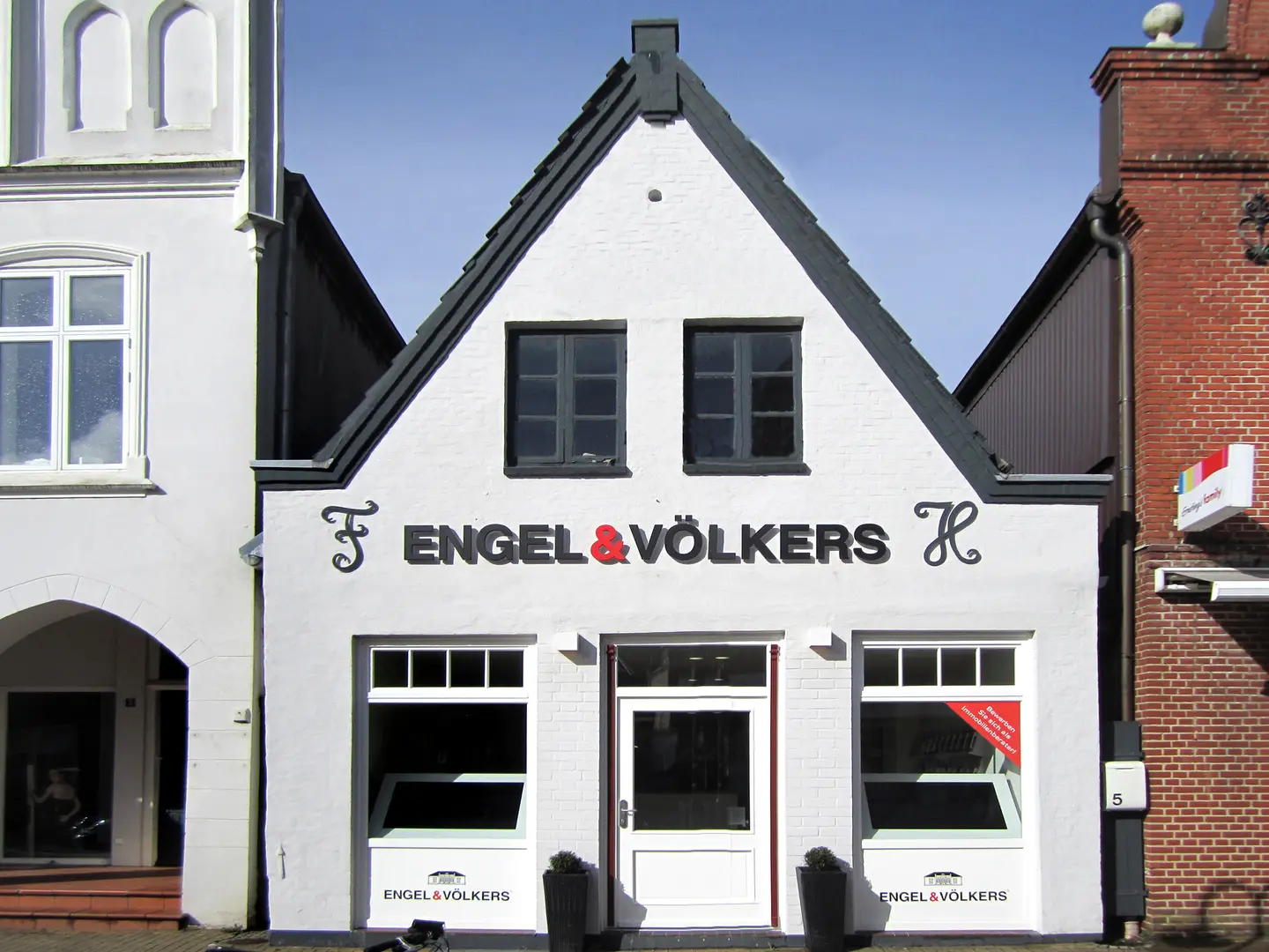 Engel & Völkers real estate office. White building with a gray roof, black lettering, and a red ampersand. Two potted plants flank the door.