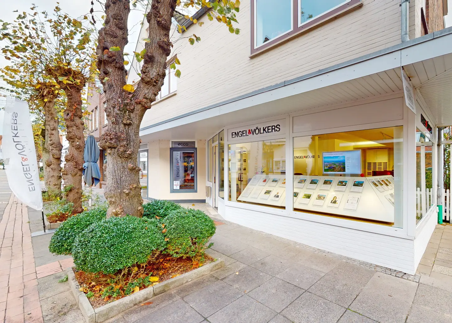 Exterior view of Engel & Völkers real estate office with display of property listings in the window. Trees and bushes line the sidewalk.