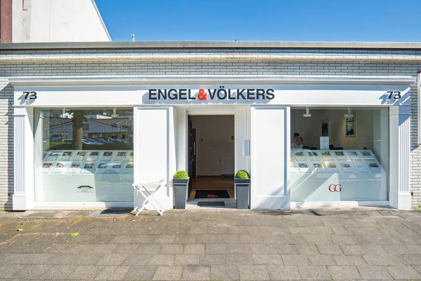 Engel & Völkers Immobilienbüro mit weißer Backsteinfassade, großen Fenstern mit Immobilieneinträgen und einer offenen Tür.
