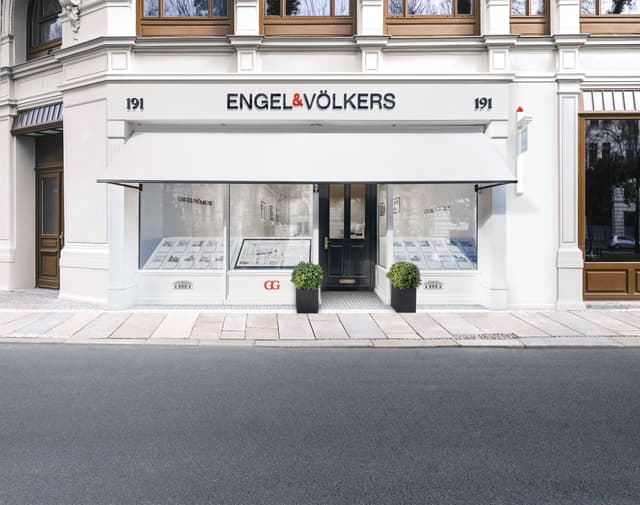 Engel & Völkers real estate office at 191, with a white awning, large windows displaying property listings, and potted plants flanking the entrance.
